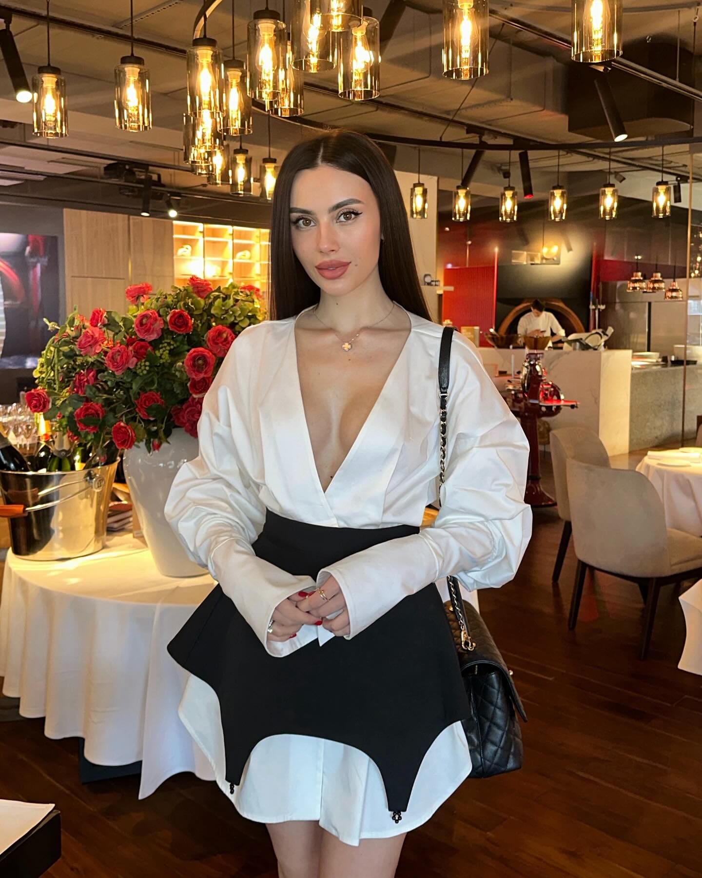 Ricky stands in a restaurant wearing a white deep-V blouse and black-and-white peplum skirt, holding a black quilted bag near red roses.