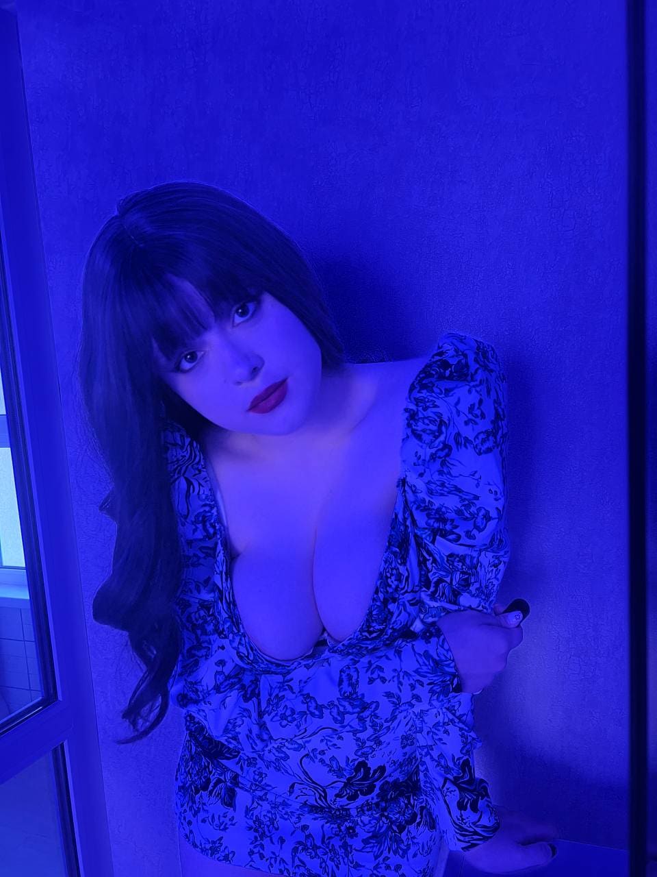 Under blue lighting, a woman with dark hair stands against a wall wearing a floral dress with a deep V-neck and puffed sleeves.