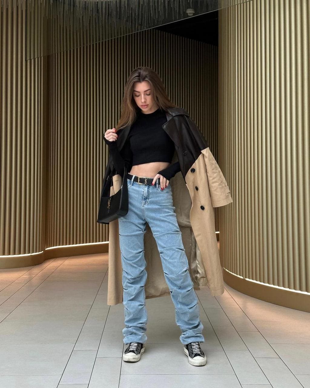 A woman stands in a tiled space with wooden slat walls, wearing a black cropped turtleneck, jeans, sneakers, and a tan coat draped over her arms.