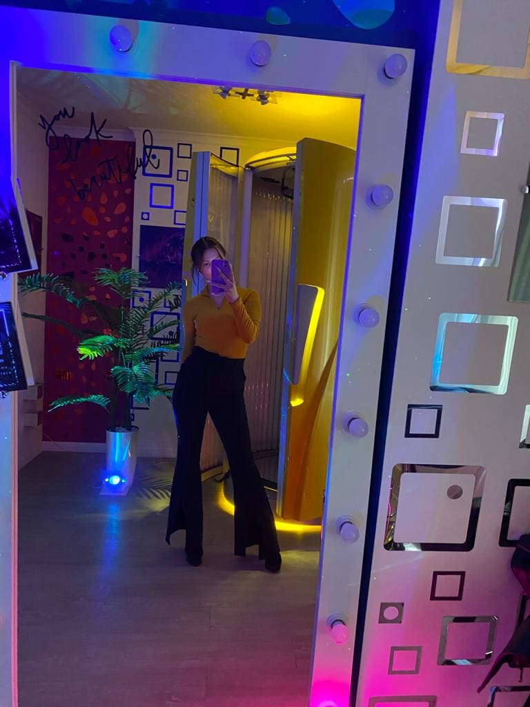 Mirror selfie in a doorway wearing a mustard top and black flared trousers, with neon lighting and a potted plant.