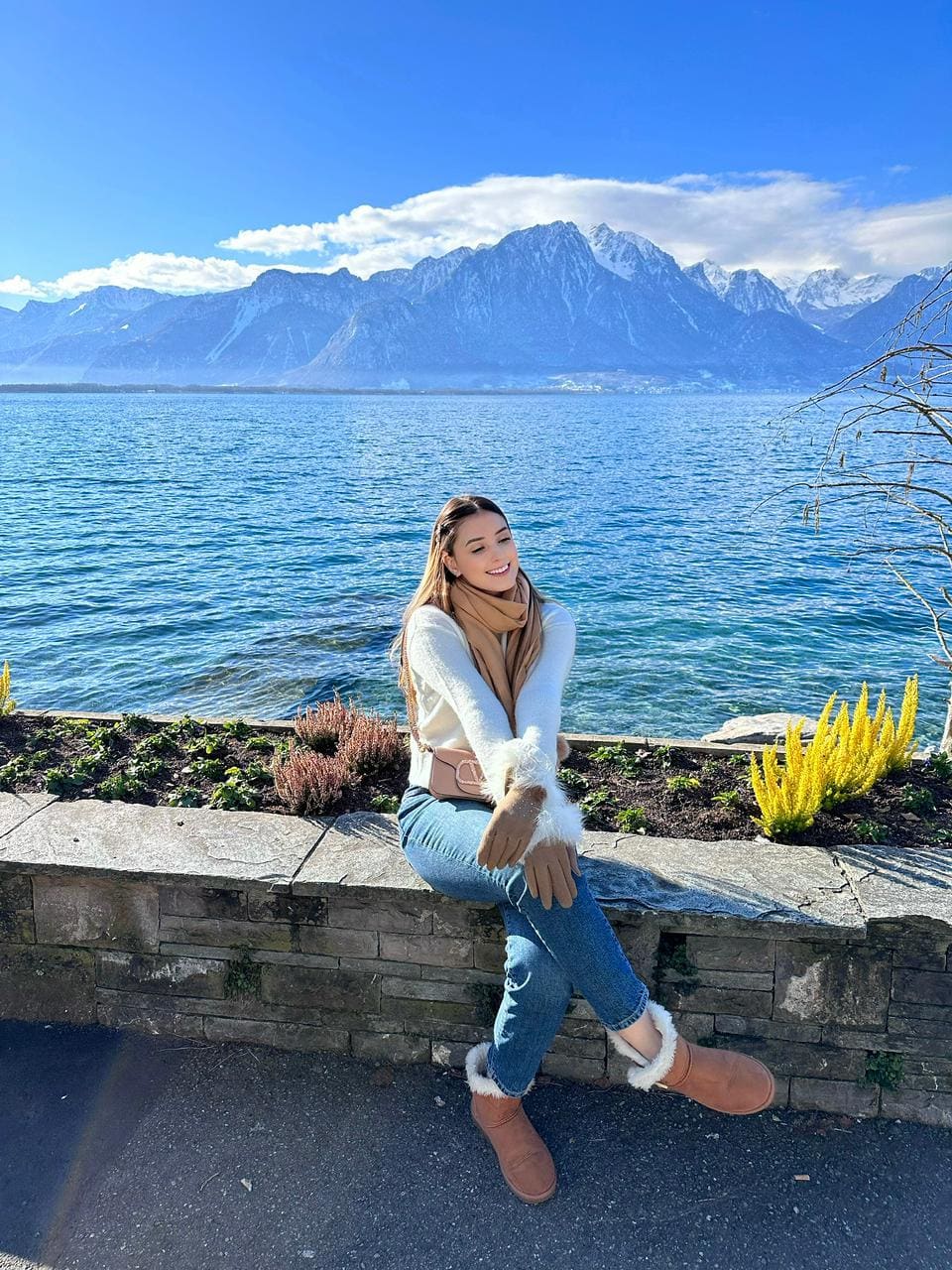 A blonde woman sits on a stone ledge by a lake in a white sweater, jeans, scarf, and fur-trimmed boots.