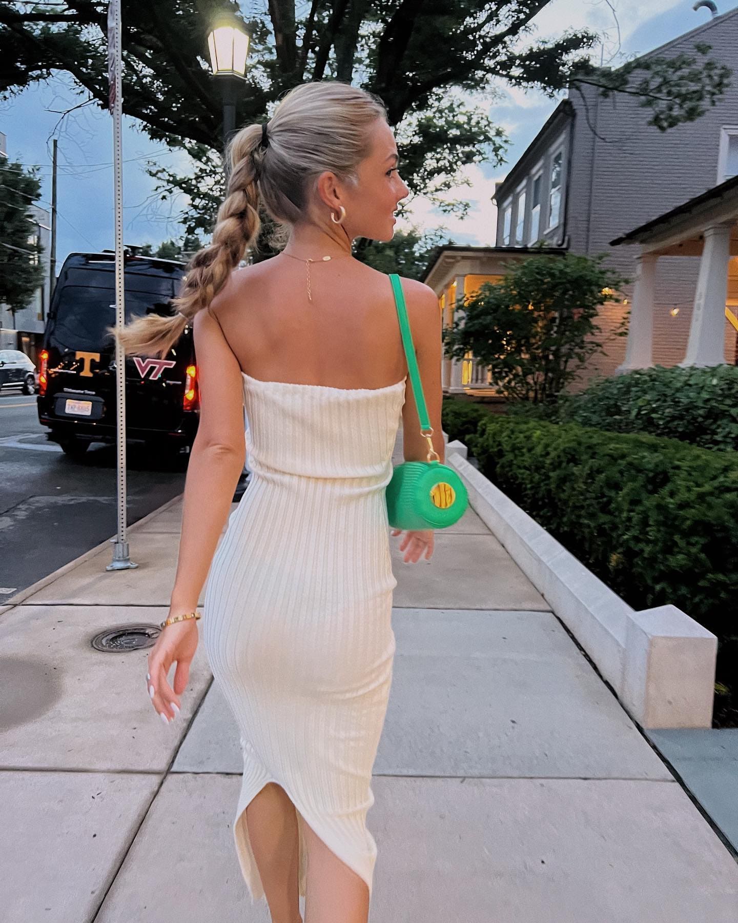 A woman with a braided ponytail is seen from behind in a white strapless dress with a slit, carrying a green handbag on a sidewalk.