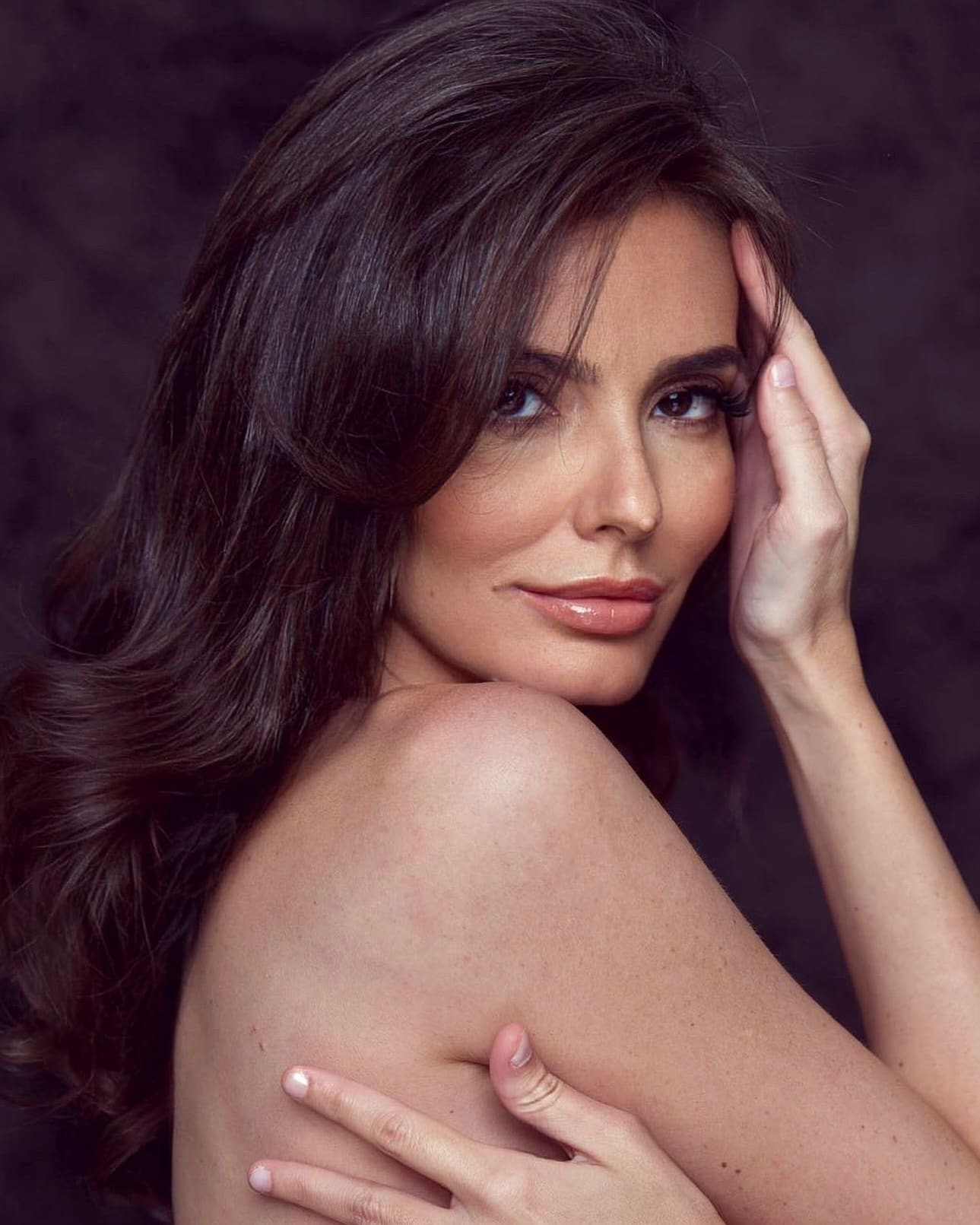 Lulu, bare-shouldered with long dark wavy hair, rests one hand on her head and the other on her back against a dark mottled backdrop.