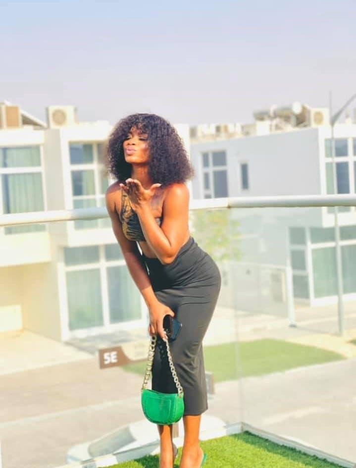 A woman with voluminous curly hair stands on a rooftop terrace wearing a dark bandeau top, long skirt, and holding a green chain-strap bag.
