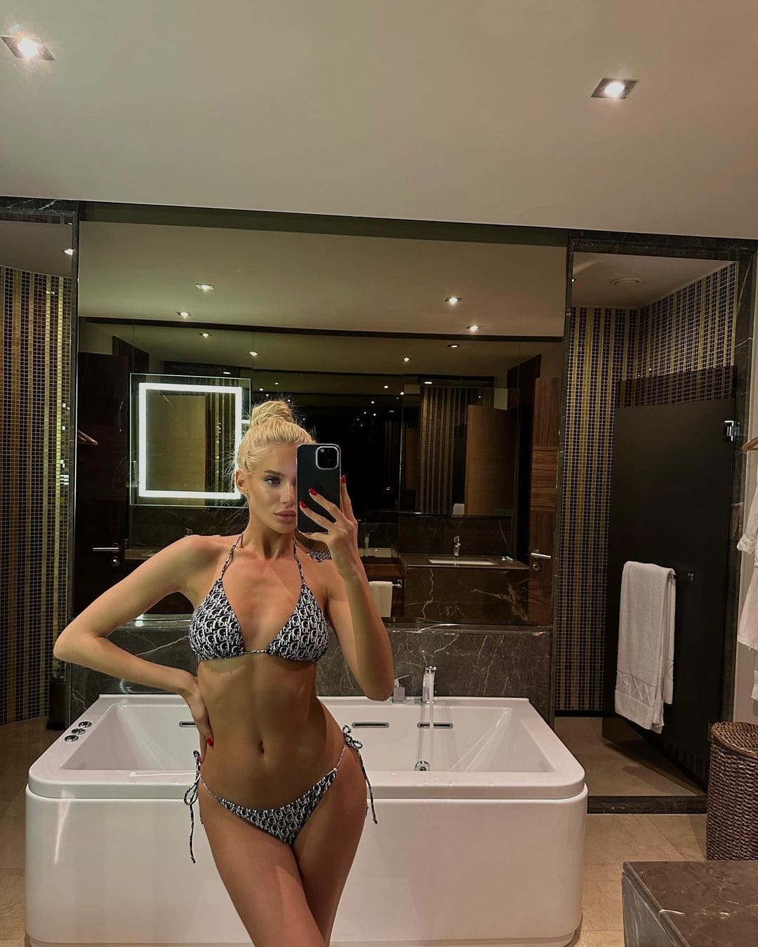 A woman with a high bun takes a mirror selfie in a bathroom, wearing a black-and-white patterned bikini in front of a white bathtub.