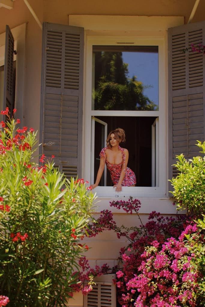 A woman in a short floral dress leans from a window with grey shutters, framed by red and pink flowering plants.