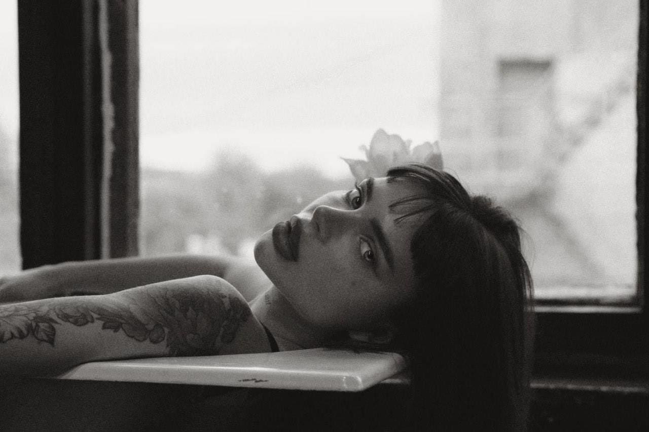 Black-and-white portrait of a woman with a floral arm tattoo lying back on a white surface, positioned in front of a large window.