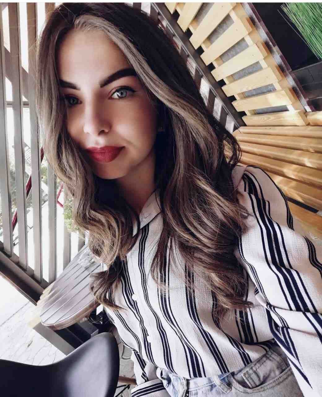 Close-up portrait in a striped white shirt and light jeans, posing in front of a wooden slatted structure and metal railing.