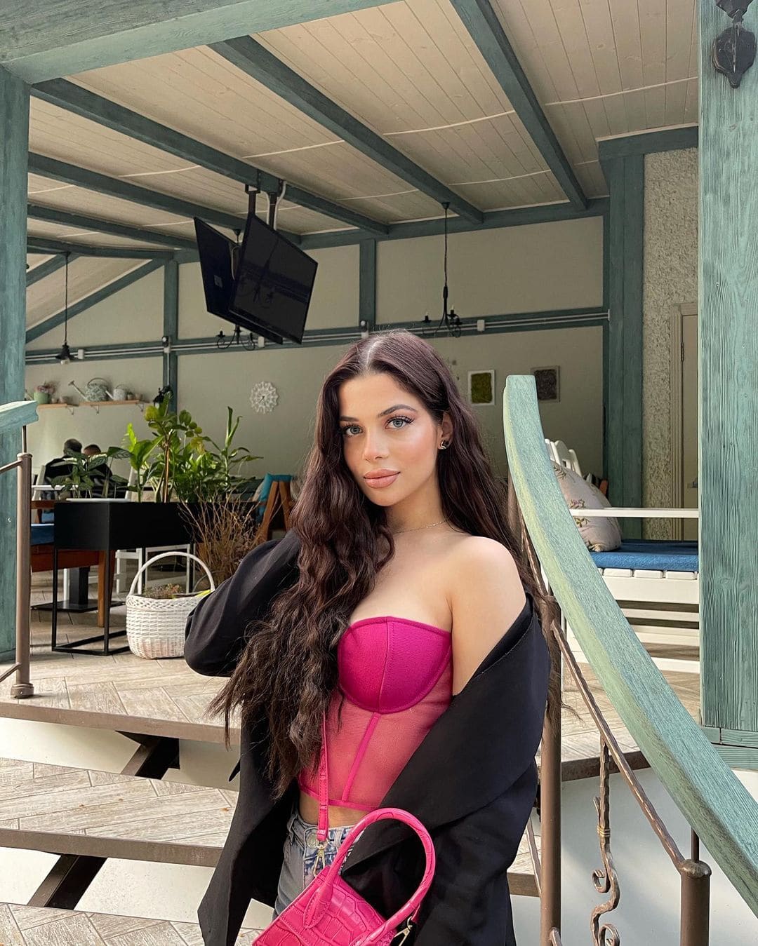 Ella stands in a covered outdoor area wearing a pink strapless bustier, a black jacket draped off one shoulder, and holding a pink handbag.