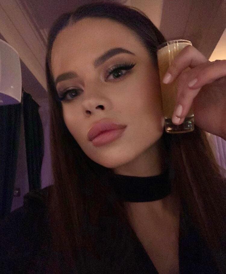 Holding a glass of amber liquid near her face in a dimly lit room with purple lighting, wearing a black choker.