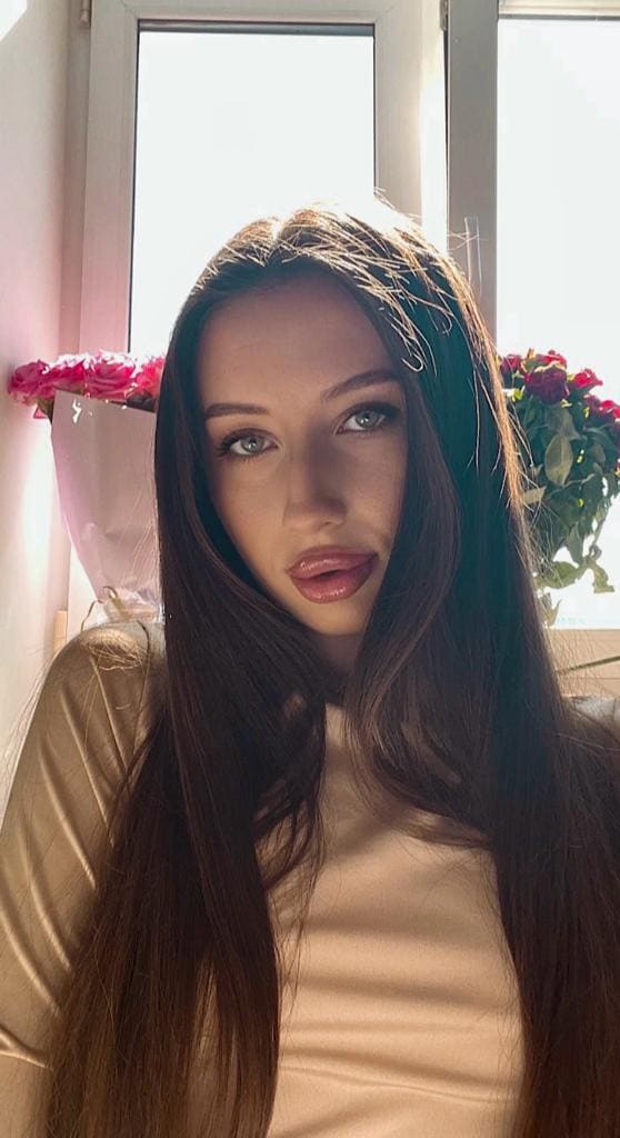 A woman in a light top sits indoors by a window, with bouquets of pink and red flowers arranged on the sill behind her.