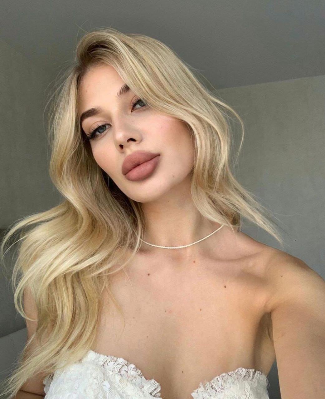 Genevieve poses in a white lace strapless top and beaded necklace against a plain gray wall, her long wavy blonde hair visible.