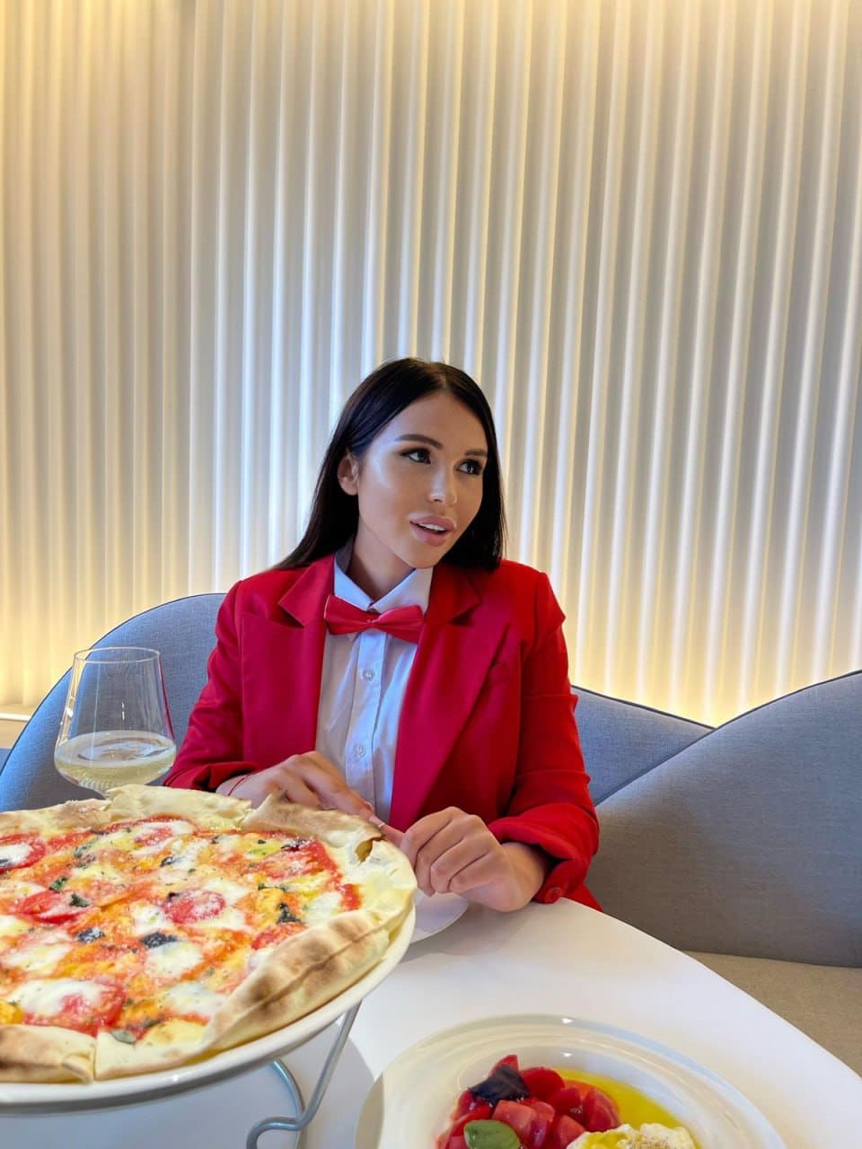 A woman in a red blazer and bow tie sits at a white table with pizza and white wine against a ribbed cream wall.