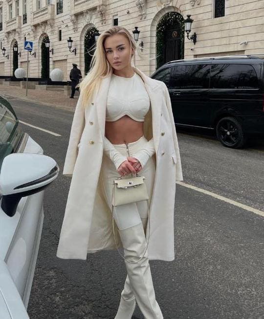 Kara stands on a city street in a long white coat over a white top and pants, holding a white handbag near parked vehicles and a stone building.