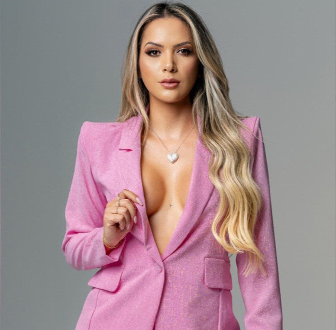 A woman with long blonde hair wears a deep-open pink blazer and heart pendant, hand near lapel, against gray.