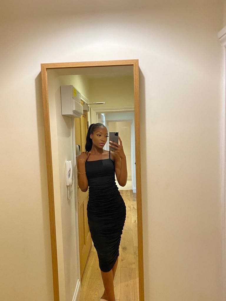 Mirror selfie of a woman in a black ruched midi dress holding a phone in a hallway with wood floors.
