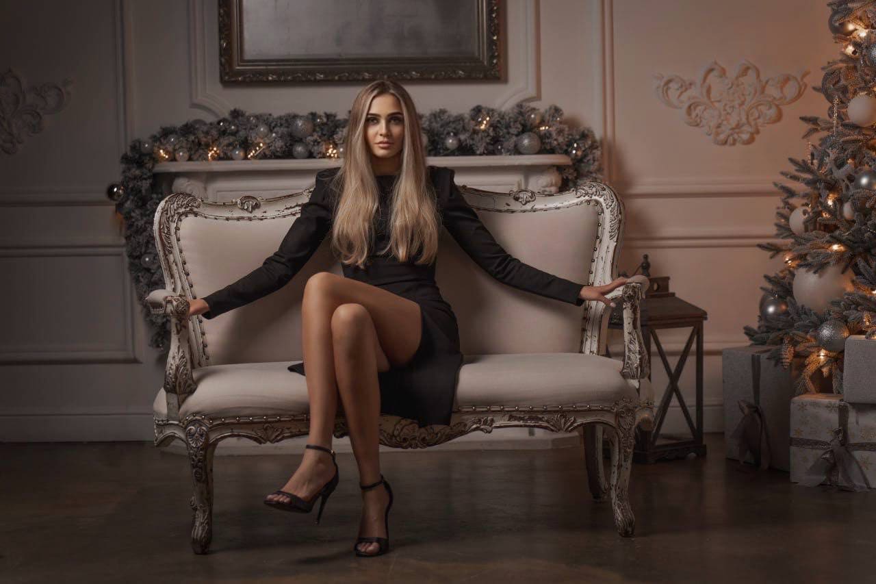 A woman sits on a white sofa in a black slit dress and heels, in a room with a garlanded mantel and Christmas tree.