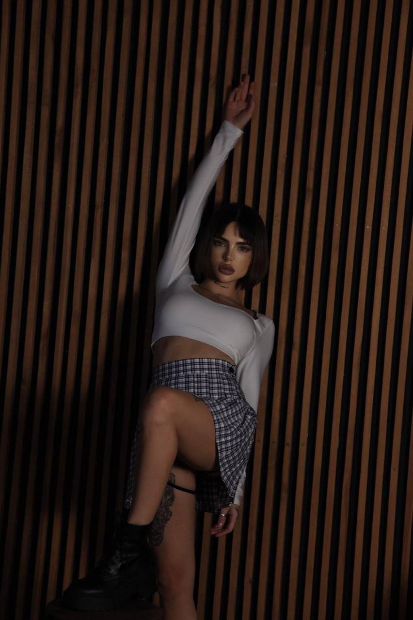 Standing against a wooden slat wall, she wears a white crop top, plaid skirt, and black boots, lifting one leg with an arm raised.