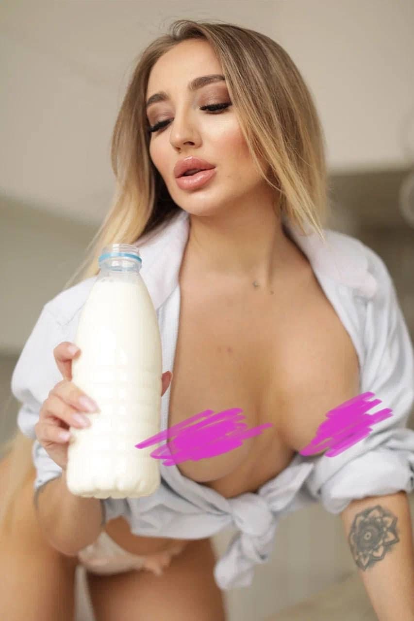 A blonde woman holds a milk bottle while wearing an open white shirt, revealing a forearm tattoo indoors.