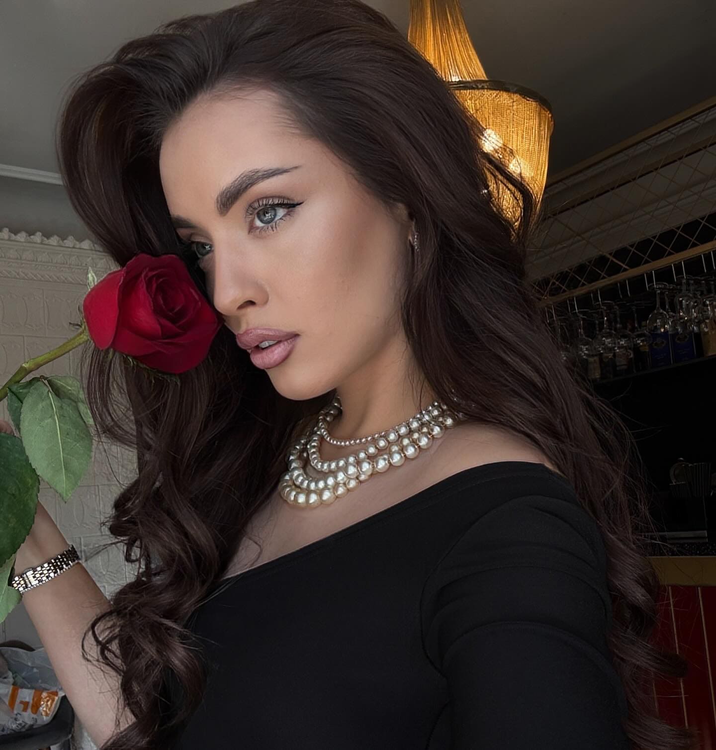 A woman in a black off-shoulder top and pearl necklace holds a red rose near her face in an indoor setting with shelves.
