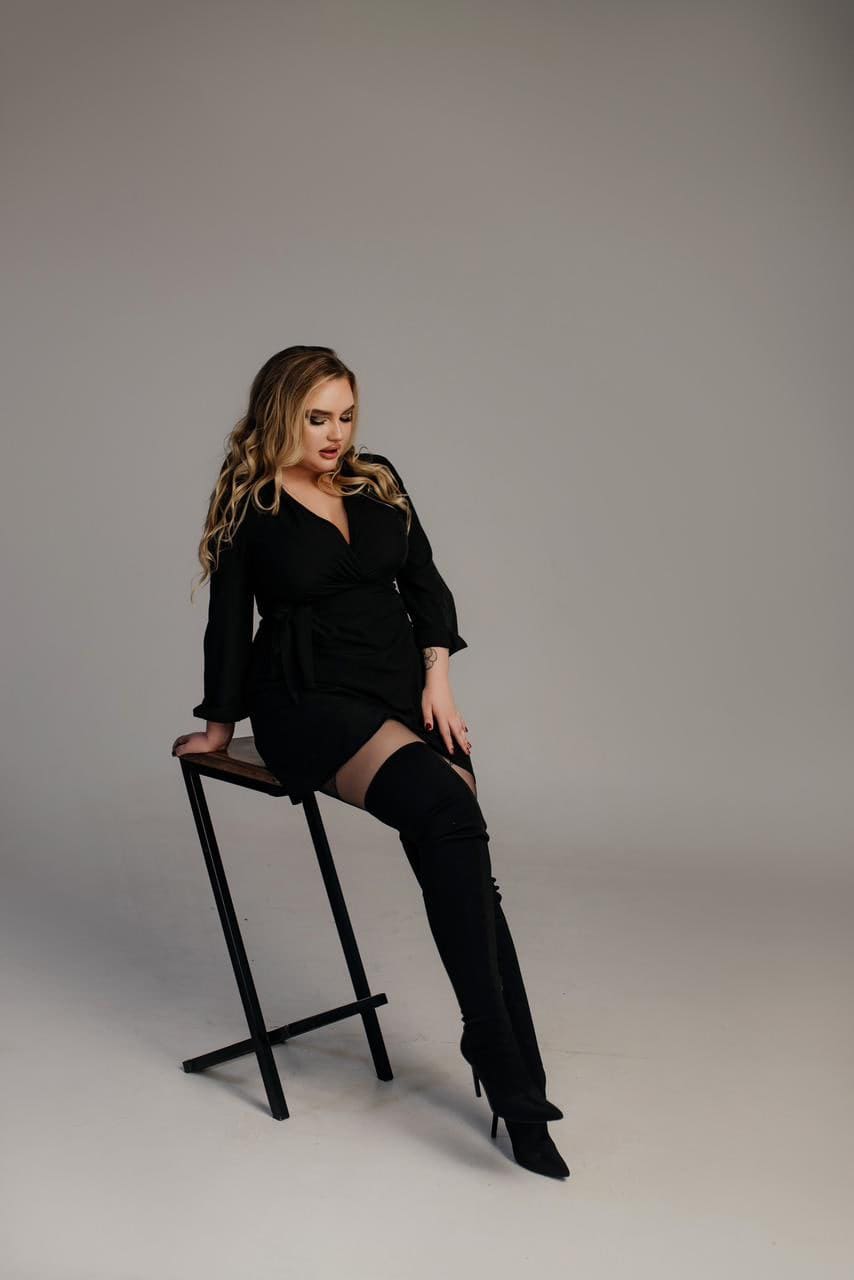 A blonde woman sits on a black stool in a black wrap dress and thigh-high boots, posing against a plain gray studio background.
