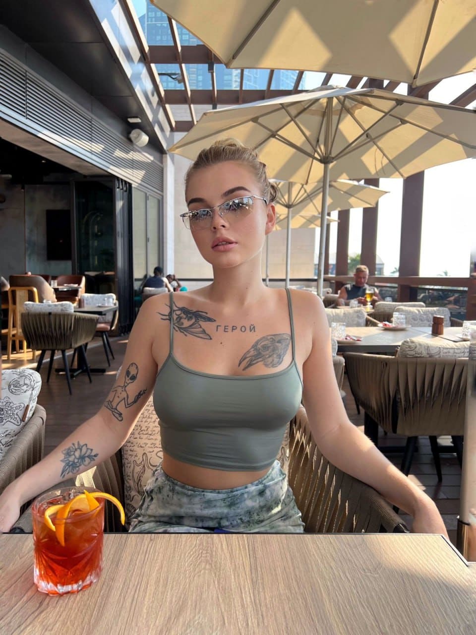 A young woman in a grey tank top and shorts sits at an outdoor table with tattoos and iced tea.