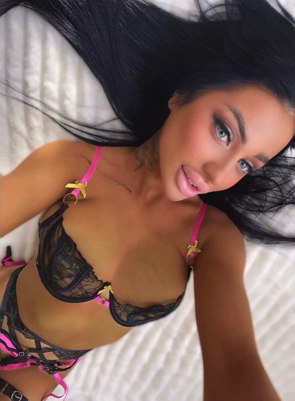A woman lies on a white bed wearing a black lace bra with pink straps and matching bottoms, looking up at the camera.