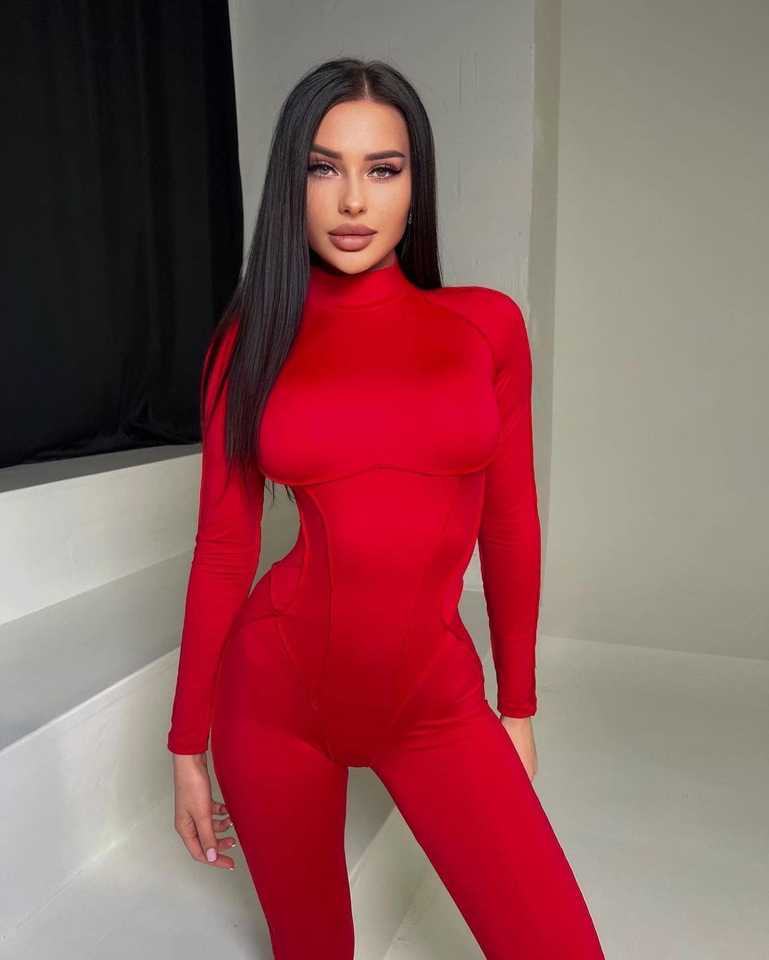A woman poses in a white room wearing a red long-sleeved turtleneck bodysuit, resting one hand on a ledge.