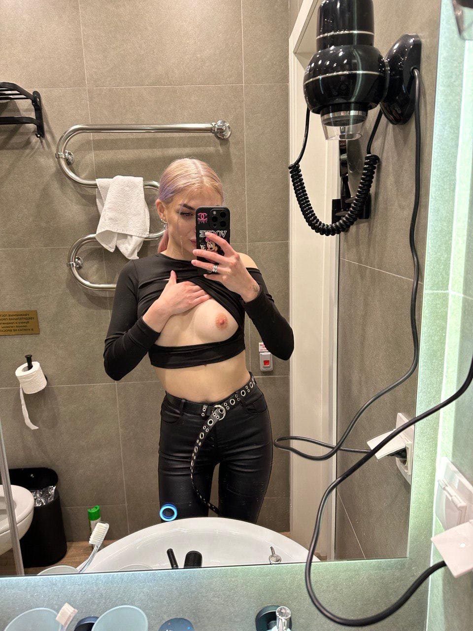 Bathroom mirror selfie in black crop top and leather pants, lifting the top with one hand while holding a phone.