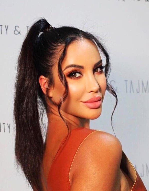 A woman with a high ponytail wears a sleeveless rust-colored top, posing with her back partially turned against a white backdrop with faint text.