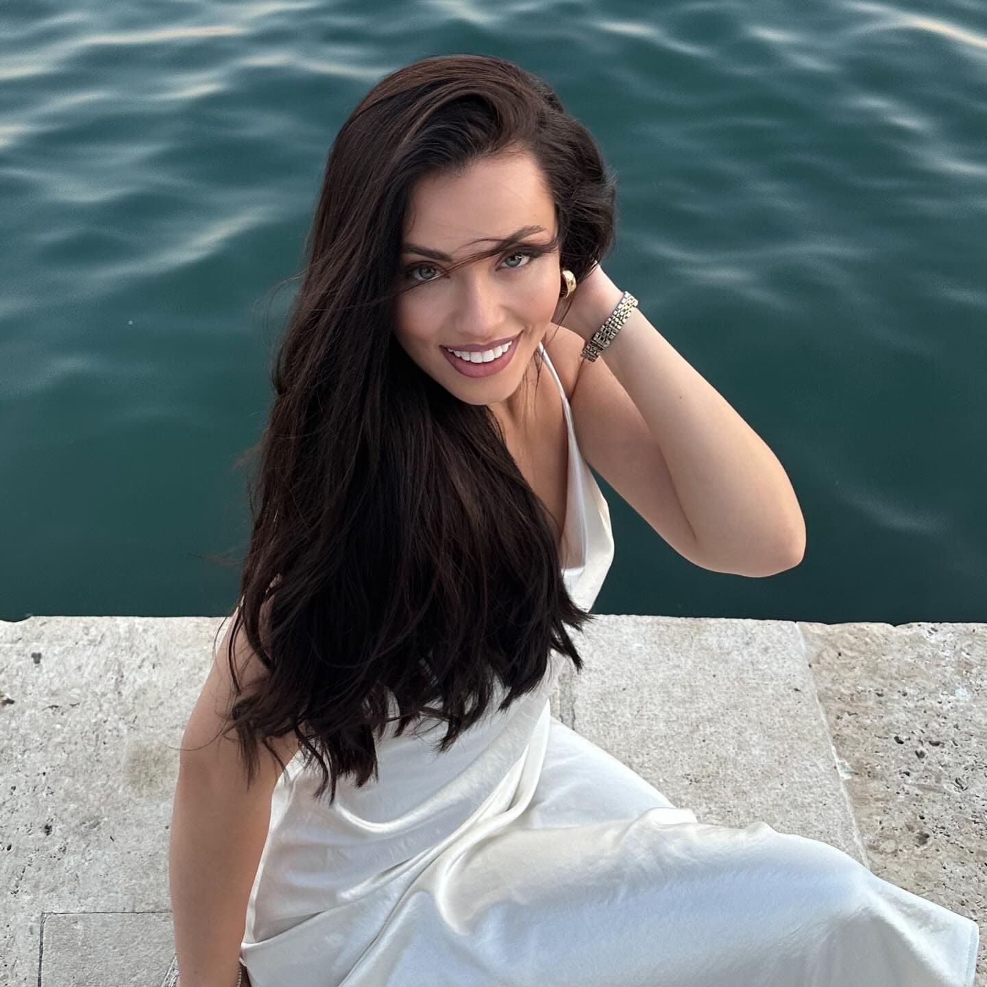 A woman sits on a stone ledge by dark water in a white satin slip dress, hand raised to her hair, wearing a bracelet.