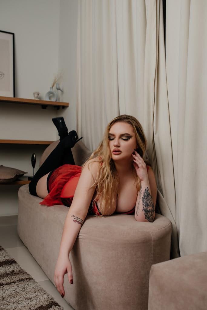 A woman with arm tattoos lies on a beige sofa in a red tulle dress, black stockings, and heeled boots, resting near her face in a bright room.