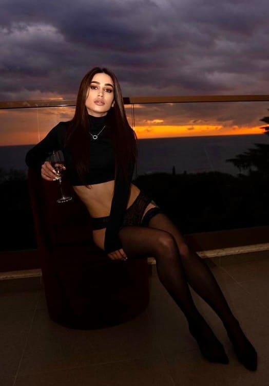 A woman in a black crop top, garter belt, and heels sits on a terrace at sunset, holding a wine glass.