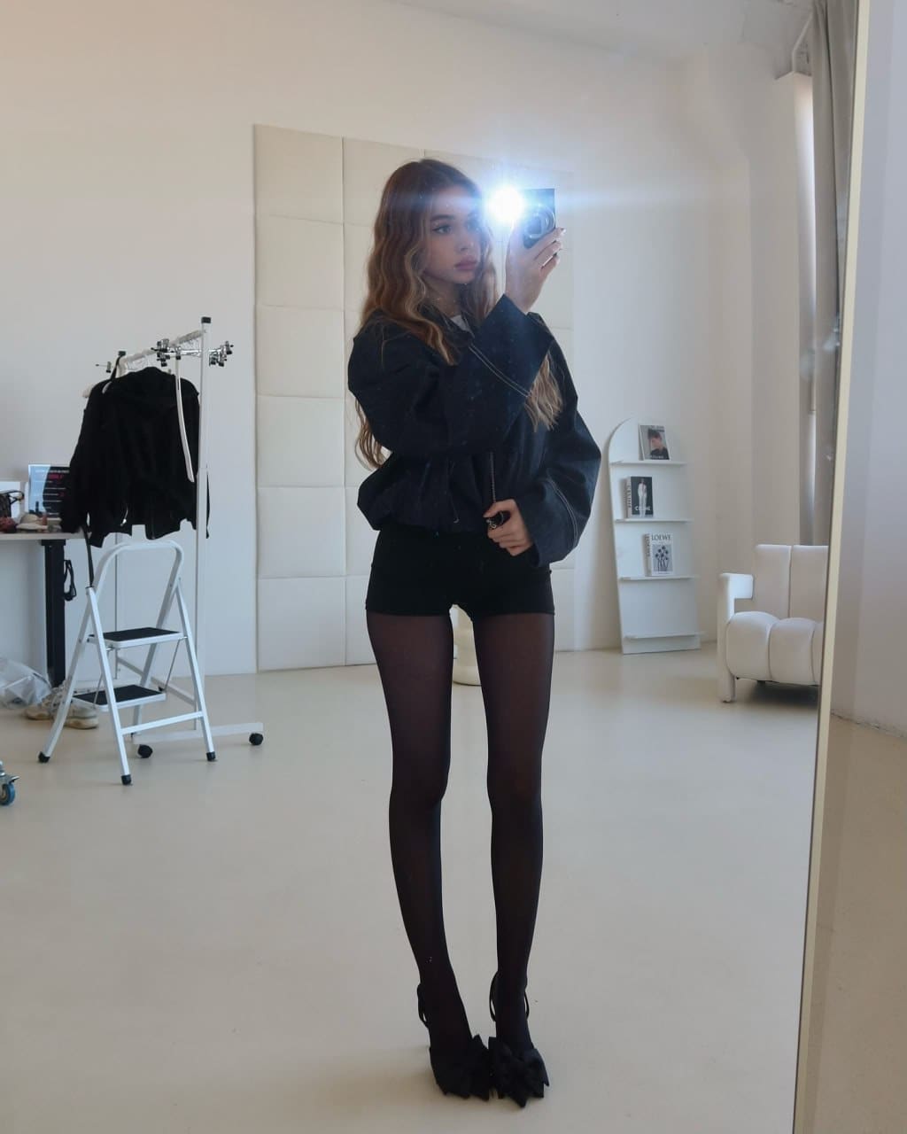 Standing in a bright white room, she takes a mirror selfie in a black jacket, shorts, tights, and heeled shoes.