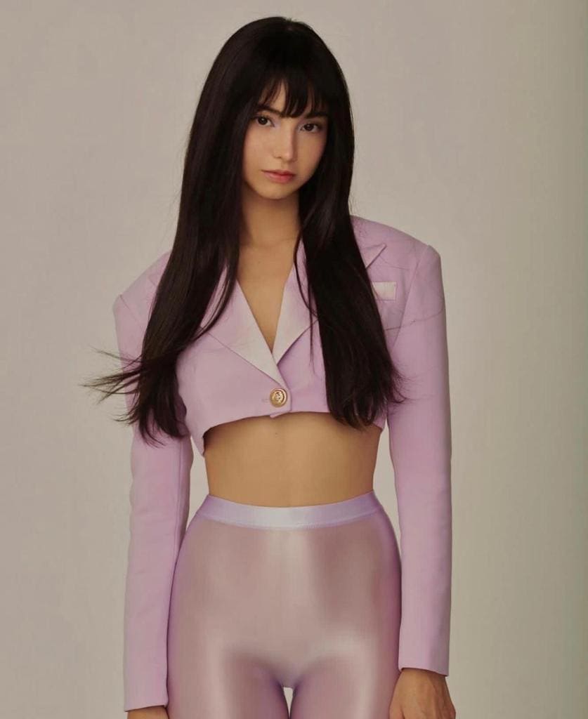 A woman with bangs stands facing the camera in a cropped light purple blazer and matching shiny leggings.