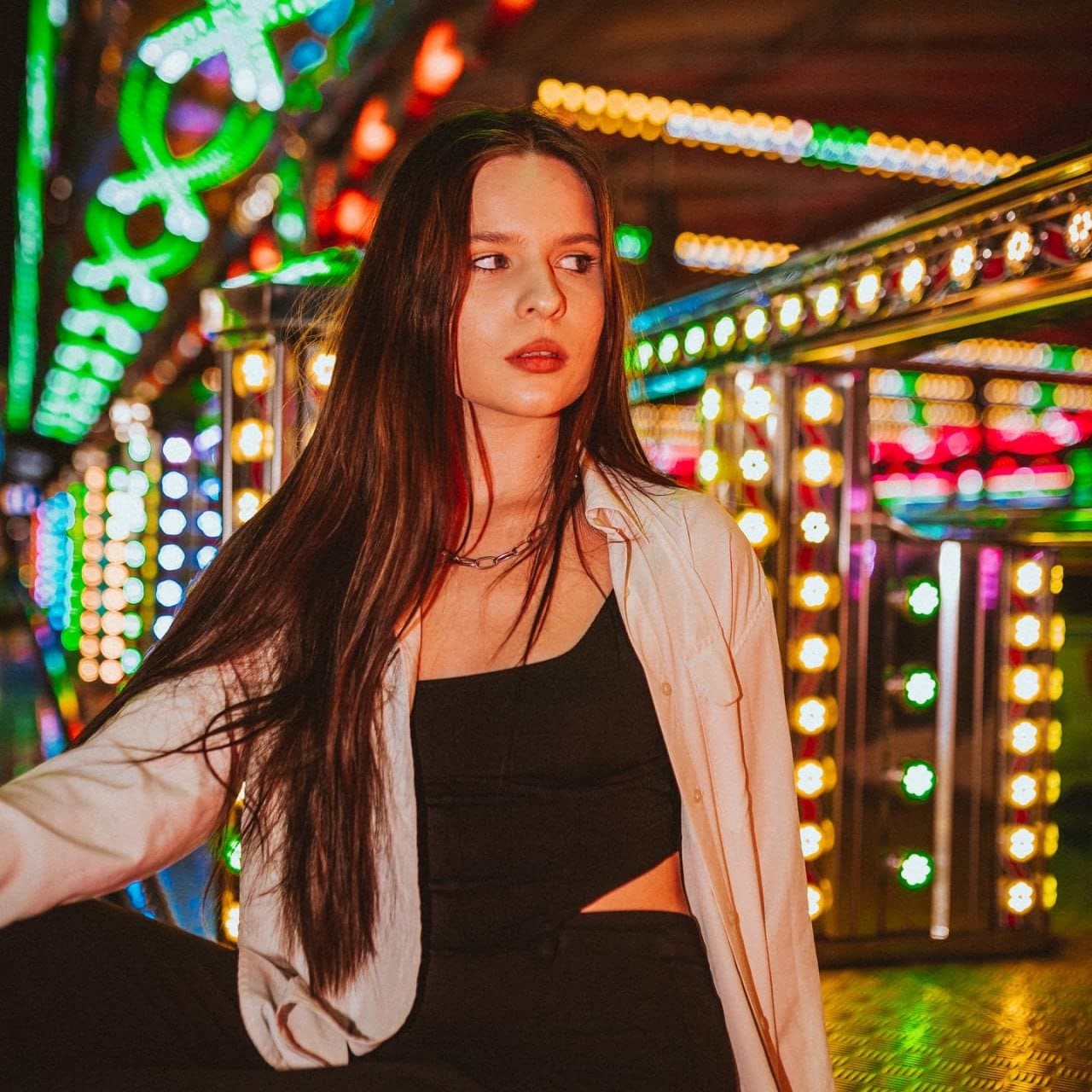 A woman sits before a lit carnival booth in a black crop top and open light shirt, looking away, chain necklace visible.
