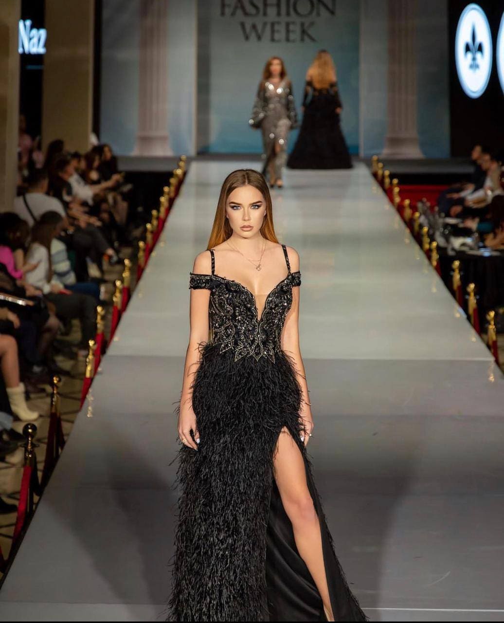 A woman walks a runway in a black off-the-shoulder gown with a sequined bodice and feathered skirt, spectators seated behind.