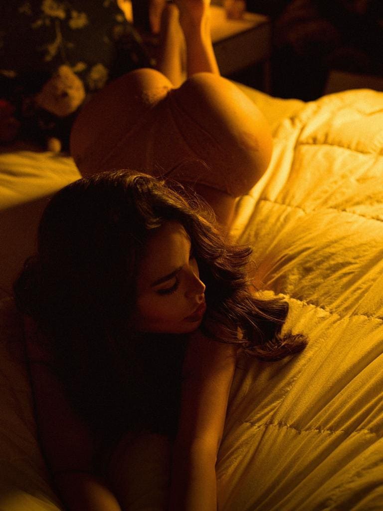 A woman lies on her side on a bed with a yellow comforter in a light-colored short dress, legs bent and raised, in a dimly lit room.