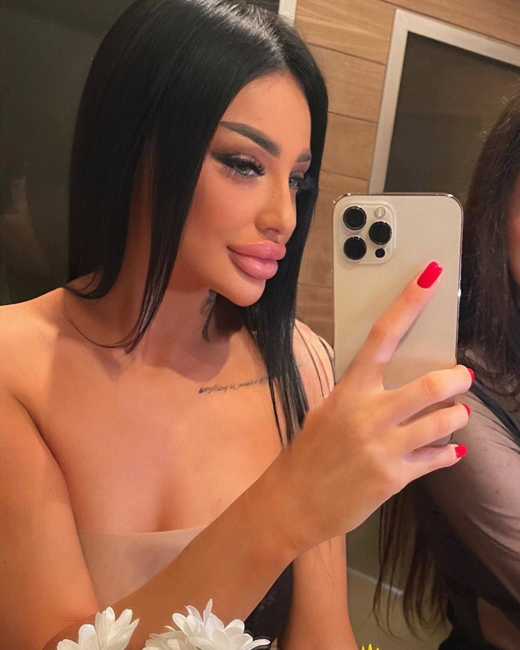 Mirror selfie of a woman in a sheer strapless top showing a collarbone tattoo, with red nails and white flowers at the bottom.