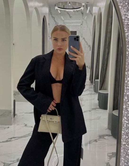 A woman takes a mirror selfie in a marble hallway, wearing an open black blazer over a black bra and trousers, holding a light-colored handbag.