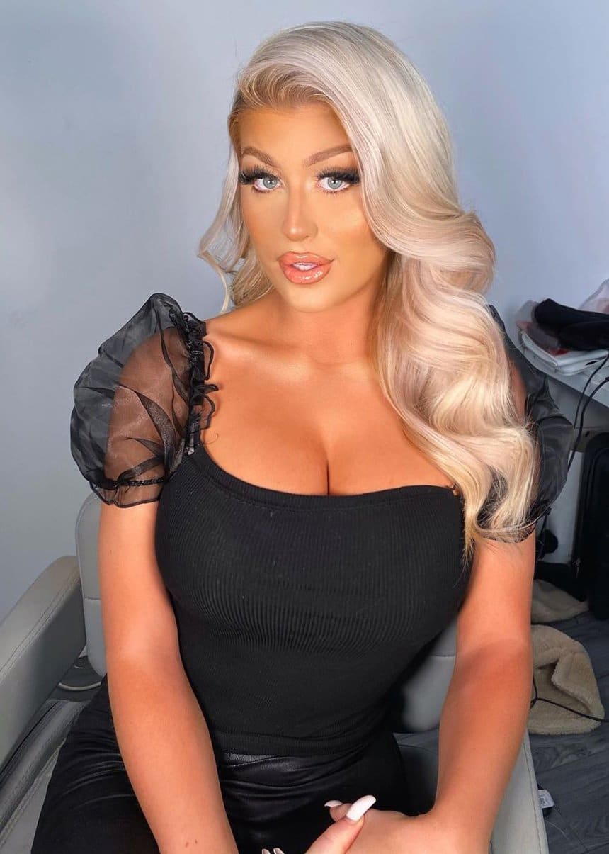 Eva sits in a light chair against a plain wall, wearing a black ribbed top with sheer puffed sleeves and a low square neckline.