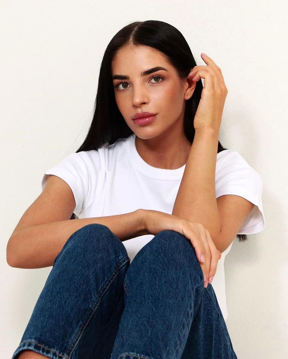 Anastasia sits with knees drawn up in a white t-shirt and blue jeans, hand in her long dark hair against a plain light wall.