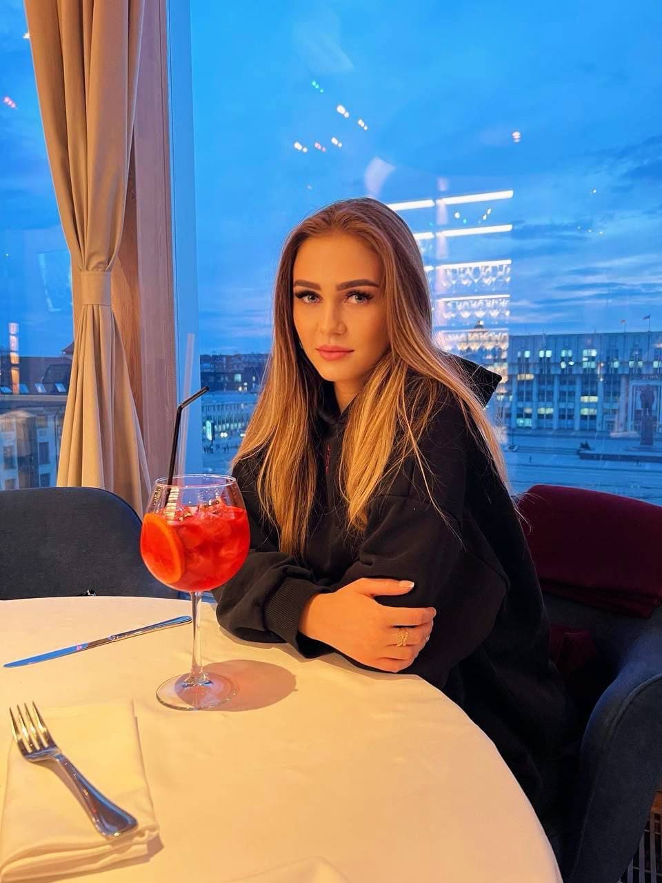 A woman in a black hooded jacket sits at a restaurant table with a drink, a city skyline visible through the window.