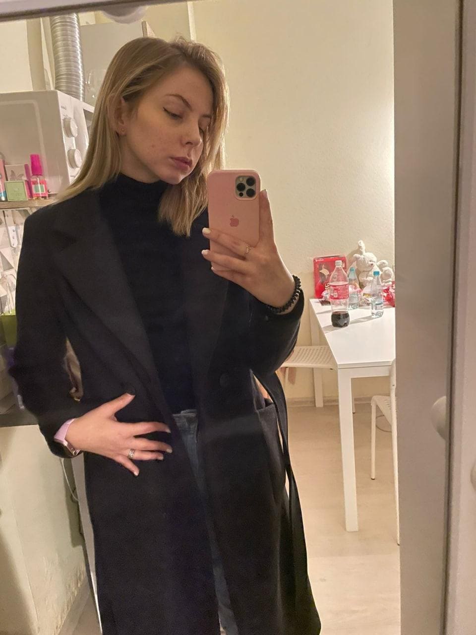 Mirror selfie of a blonde woman in a black turtleneck and long coat, standing in a room with a white table and background items.
