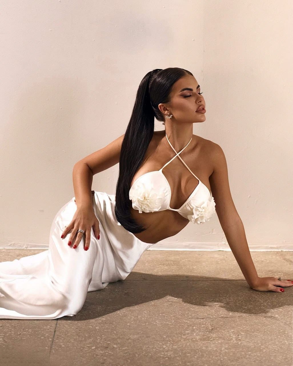 A woman with a high ponytail kneels on concrete wearing a white floral bralette and flowing skirt, head tilted back against a plain wall.