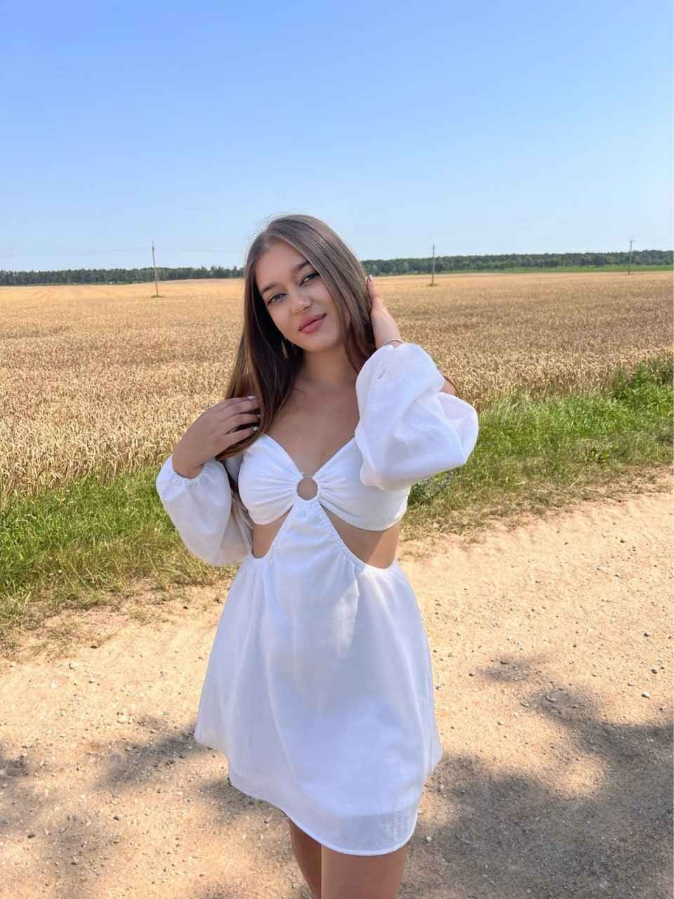 Chris stands on a dirt path by a harvested field in a white cut-out mini dress with puffed sleeves, hand raised to her hair.