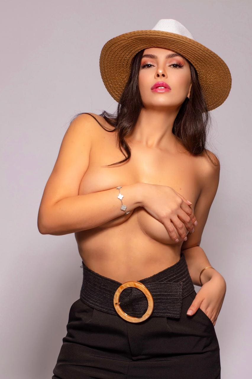 A woman in a straw hat and black trousers poses topless with an arm across her chest against a gray background.