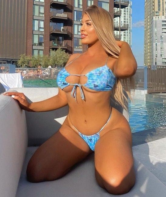 A blonde woman kneels on a ledge beside a pool in a blue patterned bikini with a cutout top, one hand touching her hair, with apartment buildings behind.