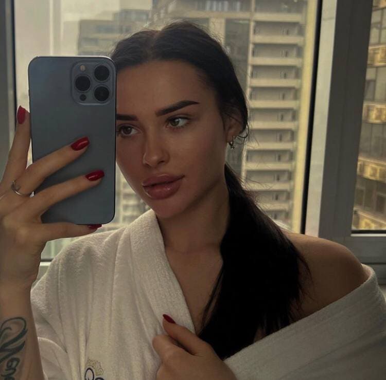 A woman with a low ponytail holds a gray phone, wearing a white towel and red nail polish, by a city-view window.