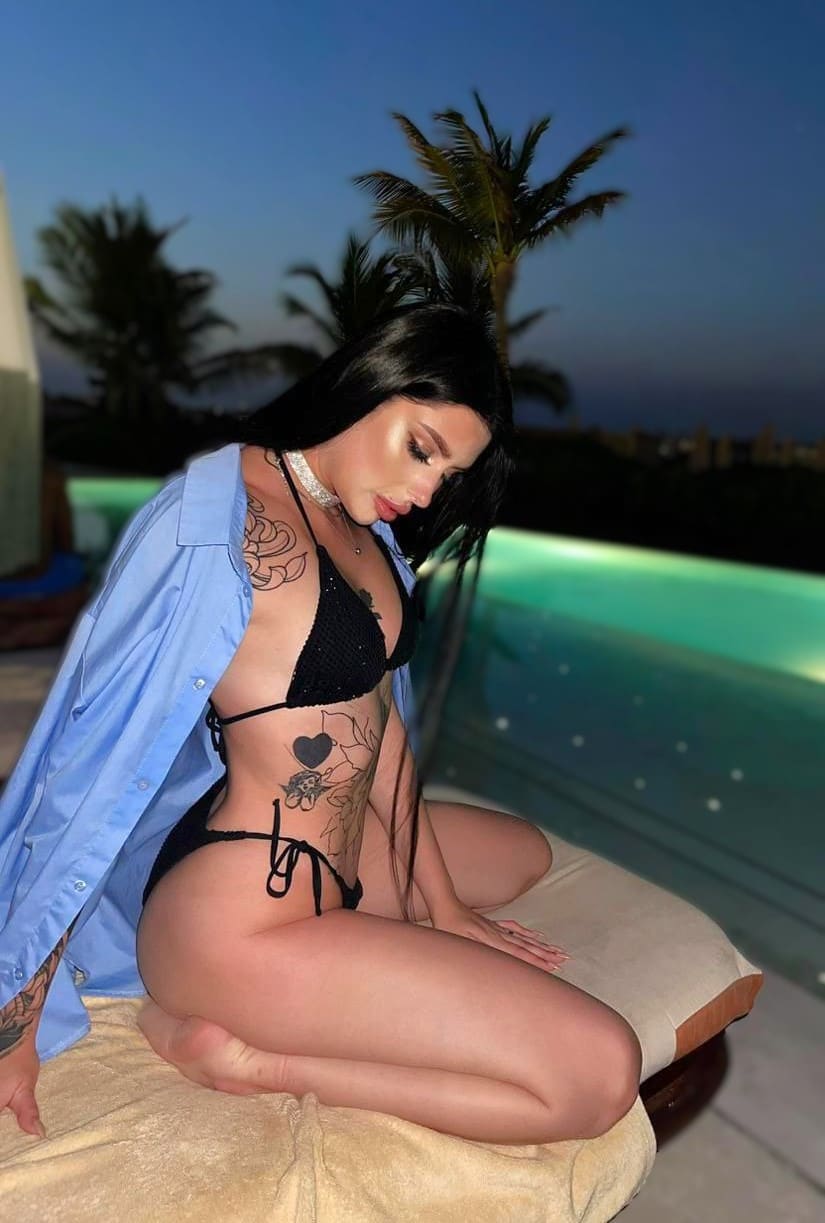 Kneeling by a lit pool at dusk, she wears a black string bikini and open blue shirt, with palm trees behind.